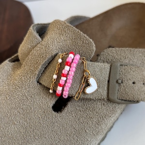Könnte beinhalten: Ein taupefarbener Wildleder-Clog mit einem dekorativen Anhänger am Riemen. Das Accessoire besteht aus goldfarbenen Ketten, einer Reihe weißer Perlen, einer Reihe abwechselnd roter und weißer Perlen, einer Reihe rosa Perlen und einem weißen Herzanhänger.