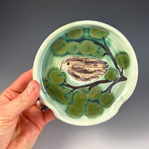 May include: A ceramic spoon rest with a bird design. The spoon rest is light green with a darker green pattern and a brown and white bird perched on a branch. The spoon rest has a curved shape with a small indentation.