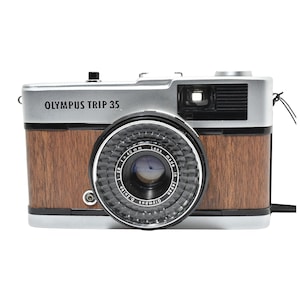 Op de afbeelding: Olympus Trip 35 camera met een zilveren bovenkant en een houten voorkant. De cameralens is zwart met zilveren details. De tekst "OLYMPUS TRIP 35" staat op de bovenkant van de camera.