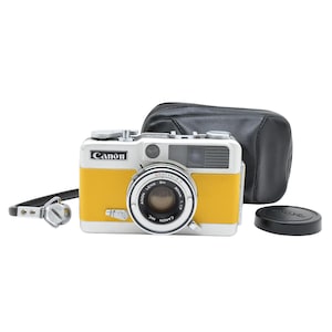 Puede incluir: Una cámara telemétrica Canon plateada y amarilla con lente central, un estuche de transporte de cuero negro, una tapa de lente negra separada y una correa para el cuello negra con un accesorio de metal. El logo de Canon aparece al frente.