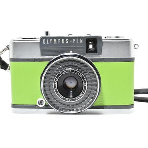 May include: A vintage Olympus-Pen camera with a silver top, a bright green body, and a black lens. The camera has a black strap and the text "OLYMPUS-PEN" is visible on the top. The lens is marked "Olympus 30mm".