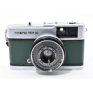 Könnte beinhalten: Olympus Trip 35 Kamera mit silbernem Oberteil, grünem Gehäuse und schwarzem Objektiv. Die Kamera hat ein klassisches Design mit einem prominenten Objektiv und verschiedenen Knöpfen. Das Objektiv trägt die Aufschrift "Lens Made in Japan".