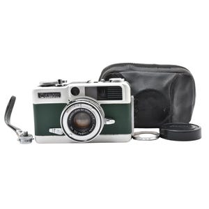Puede incluir: Una cámara Canon vintage con cuerpo verde y plateado, tapa de lente y estuche de cuero. La cámara tiene un anillo de lente plateado y una correa negra. La cámara es de diseño clásico.