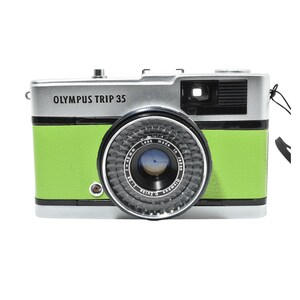 May include: Olympus Trip 35 camera with a silver top and a bright green body. The camera lens is black and gray with the text "Olympus" and "Made in Japan". The camera has a black strap attached.