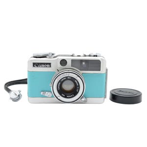 Puede incluir: Una cámara Canon de color turquesa y plata con un objetivo de 30mm 1:1.7, un visor y un botón de disparo en la parte superior. Una correa de muñeca negra está unida al lateral y una tapa de objetivo circular negra está situada a la derecha.