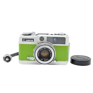 Puede incluir: Una cámara Canon plateada y verde lima con un objetivo de 30mm 1:1.7, una placa superior plateada con diales, una tapa de lente negra y una correa de muñeca negra. El cuerpo de la cámara tiene una empuñadura verde texturizada.