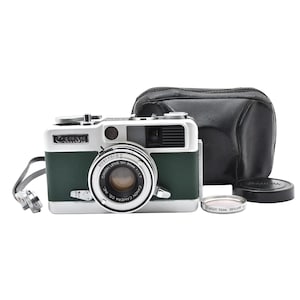 May include: A vintage Canon camera with a green and silver body, lens, and a black leather case. The camera has a lens cap and a filter. The camera strap is attached. The camera is on a white background.