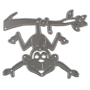 May include: A die-cut metal monkey hanging from a branch. The monkey is upside down with its arms and legs outstretched. The branch has leaves and an arrow shape on the left side. The color is a muted gray.