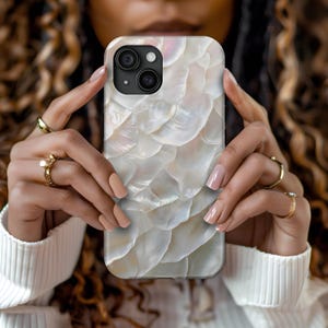 May include: A white iPhone case with a pearlescent shell pattern. The case has a black camera lens cutout.