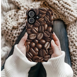 May include: A phone case featuring a close-up image of roasted coffee beans. The case is held by a person wearing a white sweater. The coffee beans are various shades of brown, with visible textures and details.