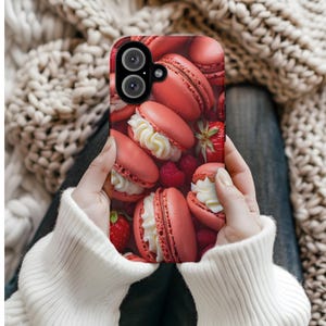 May include: A phone case featuring a close-up of pink macarons with white cream filling, raspberries, and strawberries. The case has a black camera lens and is held by a person wearing a white sweater.