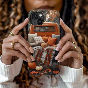 May include: A phone case featuring a fall scene with an orange pickup truck bed filled with pillows, pumpkins, and a plaid blanket. The case has a dark background with autumn leaves and string lights.