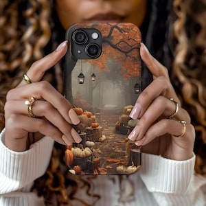 May include: A phone case featuring a fall scene with pumpkins, lanterns, and a path through a forest. The case has a dark background with orange and brown autumn leaves. The phone case is held by a person.