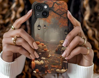 Op de afbeelding: Een telefoonhoesje met een herfsttafereel met pompoenen, lantaarns en een pad door een bos. De hoes heeft een donkere achtergrond met oranje en bruine herfstbladeren. De telefoonhoes wordt door een persoon vastgehouden.