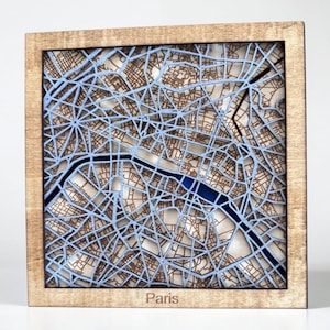 May include: Wooden wall art featuring a detailed map of Paris, France. The map is laser-cut with intricate street patterns in brown and light blue, set within a natural wood frame. The word "Paris" is at the bottom.