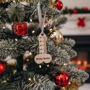 Puede incluir: Adorno de árbol de Navidad de madera con forma de regla, con el texto "Willy Ruler" y medidas en pulgadas. El adorno cuelga de una cinta gris en un árbol de Navidad decorado con adornos rojos y dorados.