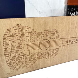 Pode incluir: Uma placa de madeira com a letra da música "Imagine" de John Lennon, com um gráfico de guitarra. O texto é gravado em uma fonte estilizada. Parcialmente visível uma garrafa de whisky.