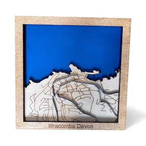 May include: A wooden framed 3D map of Ilfracombe, Devon, with a blue background representing the sea. The map is laser-cut with intricate details of the town's layout. The frame is a light natural wood color, and the text "Ilfracombe Devon" is visible.