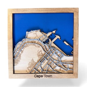 Peut inclure: Une carte en bois de Cape Town, Afrique du Sud, avec un fond bleu. La carte est découpée au laser avec des détails complexes des rues et du port de la ville. La carte est encadrée dans un cadre en bois de couleur claire, avec le nom de la ville imprimé en bas.