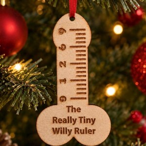 Op de afbeelding: Een fallusvormig houten ornament dat aan een rood lint hangt. Het oppervlak is voorzien van een gegraveerde liniaal met markeringen en de tekst 'The Really Tiny Willy Ruler' in het zwart. Het hout heeft een lichtbruine afwerking.