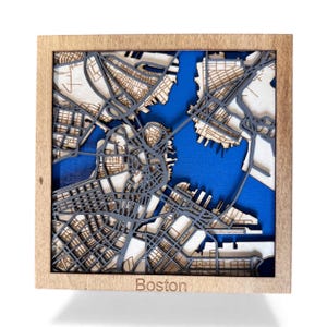Op de afbeelding: Een houten kaart van Boston, met een gelaagd ontwerp met grijze straten en waterwegen tegen een blauwe achtergrond. De kaart is ingekaderd in een lichtgekleurde houten lijst, met de stadsnaam "Boston" onderaan.