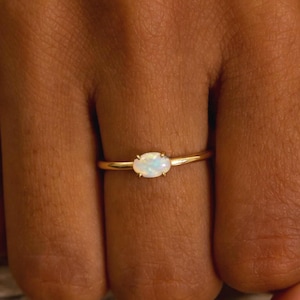 May include: A gold ring with an oval, light-colored gemstone. The ring is delicate and sits on a finger. The background is a wooden surface. The ring is a piece of jewelry.