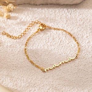May include: A delicate gold-colored bracelet with the word "mama" in cursive script. The bracelet is on a white surface with dried flowers in the background. The bracelet has an adjustable chain.