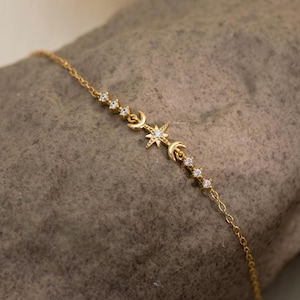 May include: A gold bracelet with a celestial design, featuring a star, crescent moons, and small, sparkling accents. The bracelet is displayed on a textured grey stone, highlighting its elegant details.