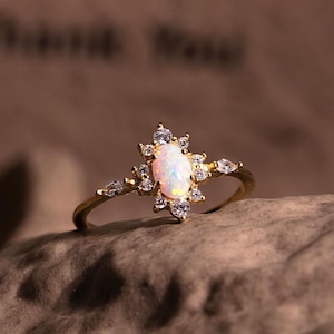 May include: A gold ring with an oval, opalescent gemstone at its centre, encircled by smaller, clear stones. The ring's slender band has additional small stones. The ring is displayed on a textured, brown surface.