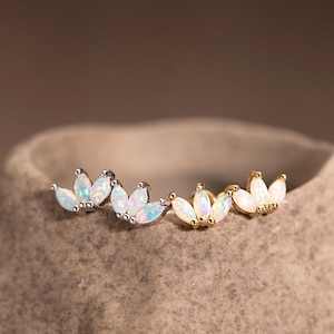 May include: A collection of opal-like stud earrings in silver and gold. Each earring features a cluster of three opal-colored stones, arranged in a floral or leaf-like design. The earrings are displayed on a textured, neutral-toned surface.