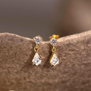 以下が含まれることがあります： クリアで輝くキュービックジルコニアの石が付いた、ゴールドカラーのドロップイヤリングのペア。各イヤリングは、上部に小さな丸い石、下部にペアシェイプの石が付いています。イヤリングは、質感のあるニュートラルなトーンの表面に表示されています。