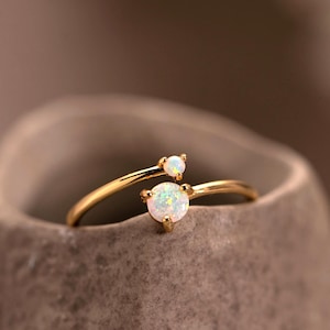 May include: A gold ring featuring two opals. The main opal is round and prominent, with a smaller opal above it. The ring is displayed against a neutral, blurred background.