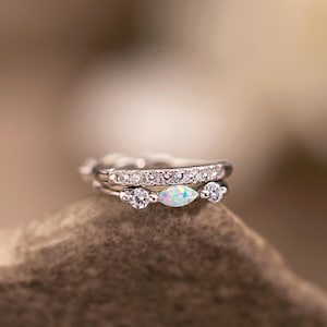 May include: Two silver rings stacked on top of each other. The top ring is encrusted with small, clear stones. The bottom ring features a central, oval-shaped opal stone, flanked by two smaller, round clear stones.