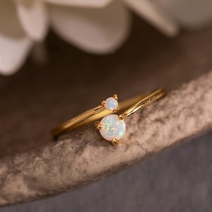 May include: A delicate gold ring featuring two opalescent gemstones. The ring has a slender band and a modern design, with one larger gemstone and a smaller accent stone. The ring is displayed on a neutral surface, with white flowers in the background.
