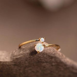May include: A gold ring with two opals. The ring's band curves elegantly, with a larger opal as the main feature and a smaller one nearby. The opals exhibit a range of colors. The ring is a minimalist design, suitable for everyday wear.