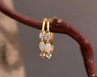 May include: A pair of gold hoop earrings with a delicate design. Each earring features a small bow-shaped accent with clear stones and a dangling cluster of white opal-like stones. The earrings are displayed on a brown twig against a blurred background.