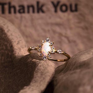 May include: A gold ring with a central oval-shaped opal stone, surrounded by smaller clear stones. The ring is set against a textured brown background, with the words "Thank You" visible in the background.