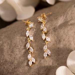 May include: A pair of gold-colored dangle earrings with a leaf-like design. Each earring features a series of clear, sparkling stones set along a delicate chain. The earrings are displayed on a gray stone, with white flowers in the background.