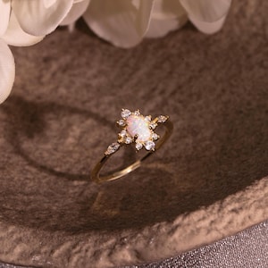 Peut inclure: Une bague en or avec une pierre d'opale ovale centrale, entourée de petits gemmes transparentes. La bague a un anneau délicat et est posée sur une surface brune texturée. Il s'agit d'un bijou.