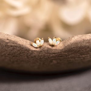 May include: A pair of gold stud earrings, each featuring three white opal-like stones arranged in a floral design. The earrings are displayed on a textured, neutral-toned surface, with a blurred background.