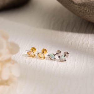 May include: Four pairs of stud earrings. Two pairs are gold-toned with opalescent planet-shaped designs. The other two pairs are silver-toned with opalescent planet-shaped designs. The earrings are displayed on a white surface.
