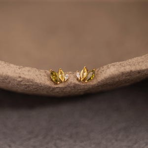 Pode incluir: Um par de brincos dourados, cada um com três pedras preciosas em forma de marquesa em tons de verde, amarelo e branco. Os brincos são exibidos em uma superfície neutra, mostrando seu design delicado e variações de cores.