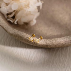 May include: Two gold stud earrings, each featuring an oval, opalescent stone. The earrings are set in a gold bezel and have a post and backing. They are presented on a neutral surface, with a blurred floral arrangement in the background.