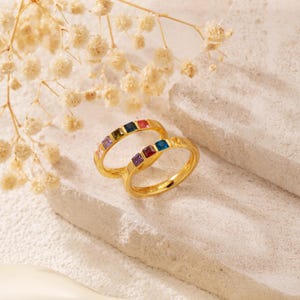 May include: Two gold-colored rings with a row of colorful square gemstones. The gemstones are in various colors, including purple, red, blue, and green. The rings are displayed on a light-colored surface with dried flowers in the background.