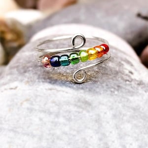 May include: A silver-toned wire ring with a rainbow of small, colorful beads. The beads are arranged in a row, transitioning from pink to red, orange, yellow, green, blue, and purple. The ring has a spiral design on each end.