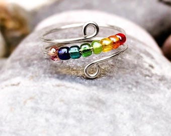 May include: A silver-toned wire ring with a rainbow of small, colorful beads. The beads are arranged in a row, transitioning from pink to red, orange, yellow, green, blue, and purple. The ring has a spiral design on each end.