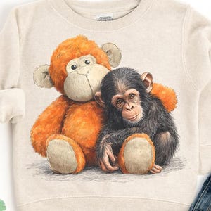 May include: A beige sweatshirt featuring a detailed illustration of a plush orange monkey hugging a black chimpanzee. The design is centered on the front of the sweatshirt. The bottom right corner of the image shows a pair of blue denim jeans.