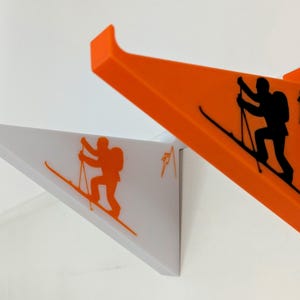 Puede incluir: Dos ganchos de pared para esquís, uno blanco y otro naranja, con una silueta de un esquiador en cada uno. Los ganchos tienen forma de esquí con un esquiador en la parte superior.