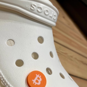 May include: White Crocs shoe with an orange and white Bitcoin charm attached to the strap.