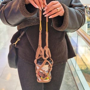 Puede incluir: Un portavasos de café tejido en color beige con una correa dorada, que sostiene una taza de café desechable. La taza tiene una tapa negra y un diseño colorido de escena de montaña. El soporte está suspendido.